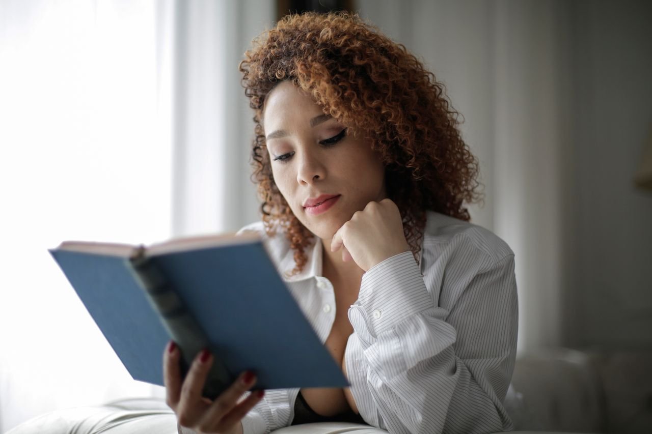 Tender curly feamle in casual white shirt reading book while resting on comfortable sofa with hand on chin in modern light room