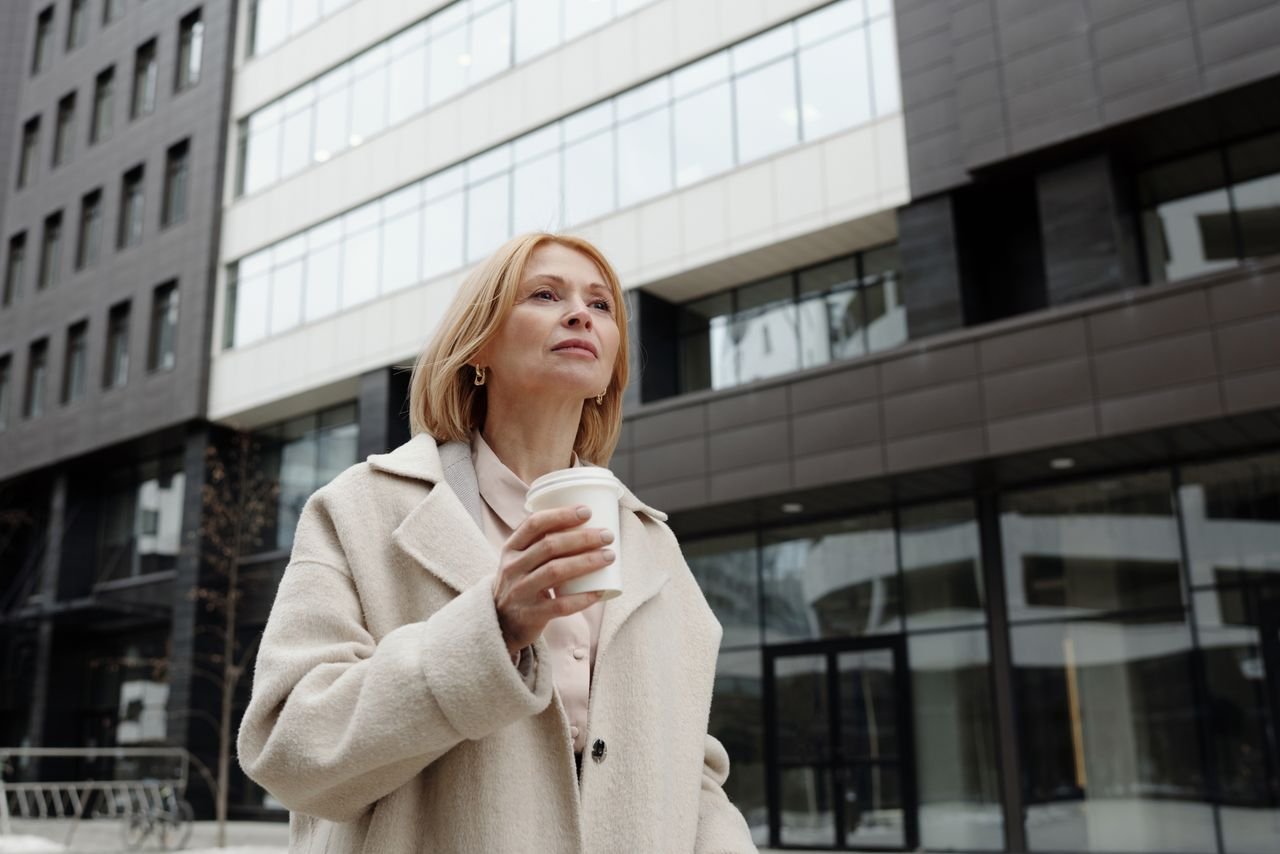 Elegant woman in beige coat holding coffee cup outside modern building.