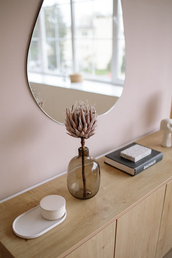 Modern interior with a wooden table, decorative mirror, and vase with dried protea flower.
