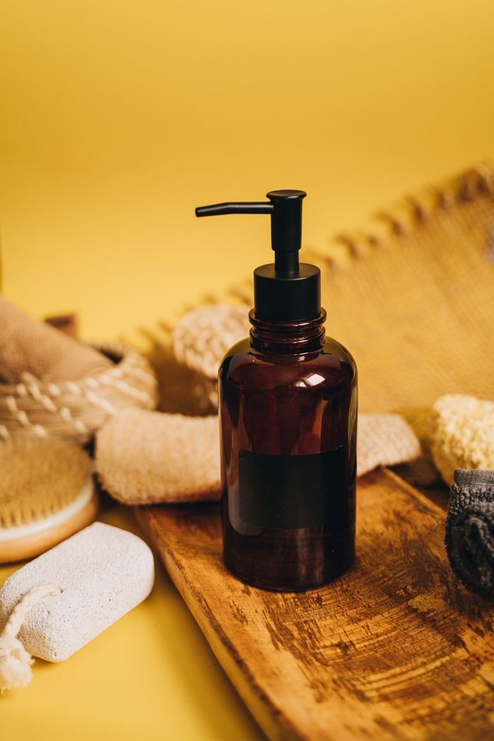 Amber pump bottle surrounded by bath accessories on a wooden tray against a warm yellow background.